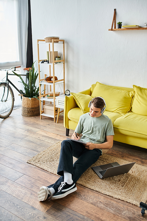 cheerful young student with headphones and laptop on floor near yellow couch writing in note