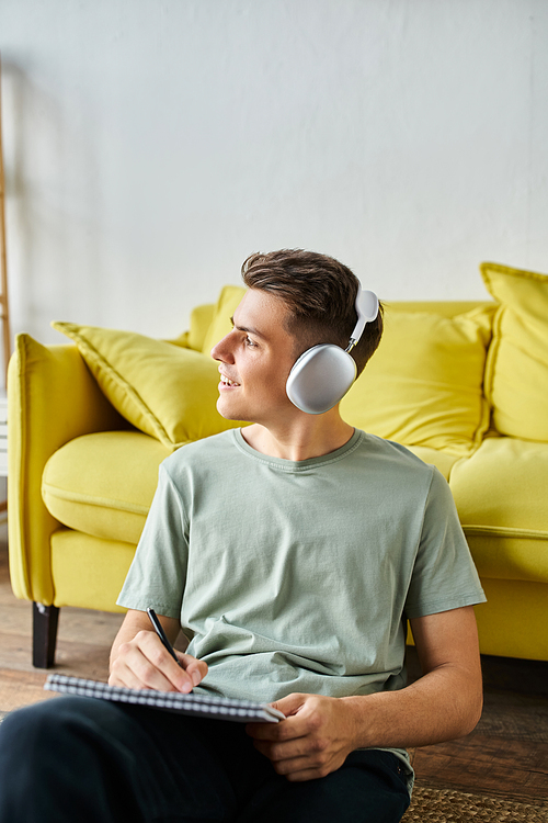 attractive man with headphones on floor near yellow couch writing in note and looking to window