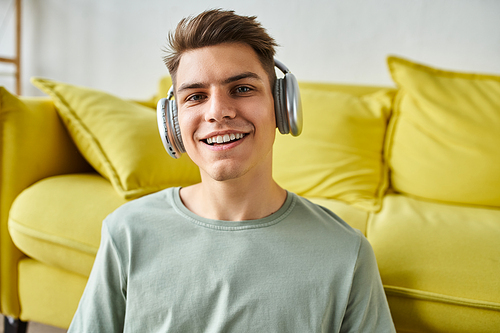 portrait of smiling charming young man in his 20s with headphones sitting near yellow couch