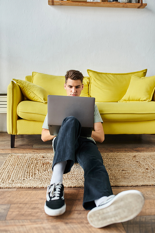 focused student in his 20s on floor near yellow couch at home networking in laptop