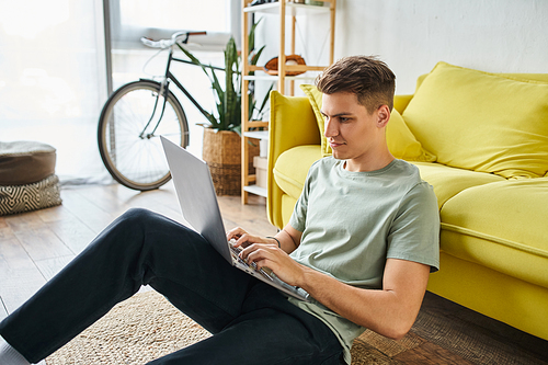 attractive student in 20s on floor near yellow couch at home networking in laptop sideways