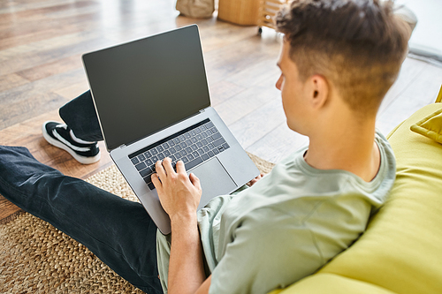 charming young man on floor near yellow couch at home networking in laptop from behind back