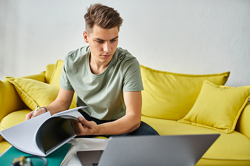 young man in yellow couch concentration doing coursework with notes and laptop on coffee table