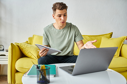 confused young student in yellow couch at home doing coursework in notes and laptop on coffee table