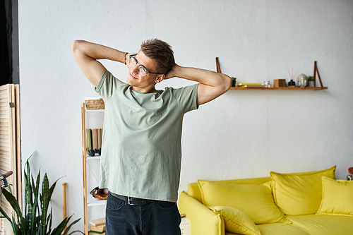charming young man with brown hair and vision glasses in living room stretching arms behind head