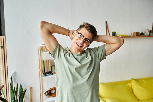 attractive guy with brown hair and vision glasses in living room putting arms behind head to camera