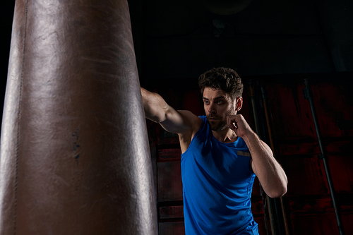 vigorous man in blue tank top boxing punching bag while training in darkness on night street
