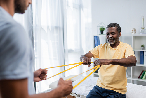 blurred rehabilitologist and african american patient doing exercise with elastics