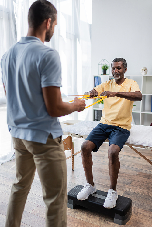 african american man doing rehabilitation exercise with elastics and blurred physical therapist