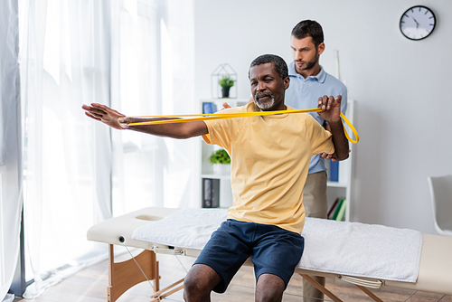 middle aged african american man sitting on massage table and training with elastics near rehabilitologist