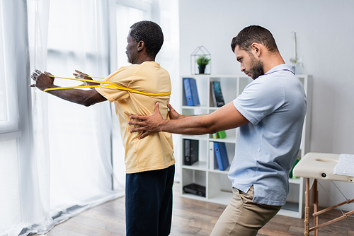 young trainer supporting mature african american man working out with elastics