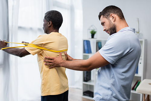 trainer assisting african american man working out with rubber band in rehabilitation center