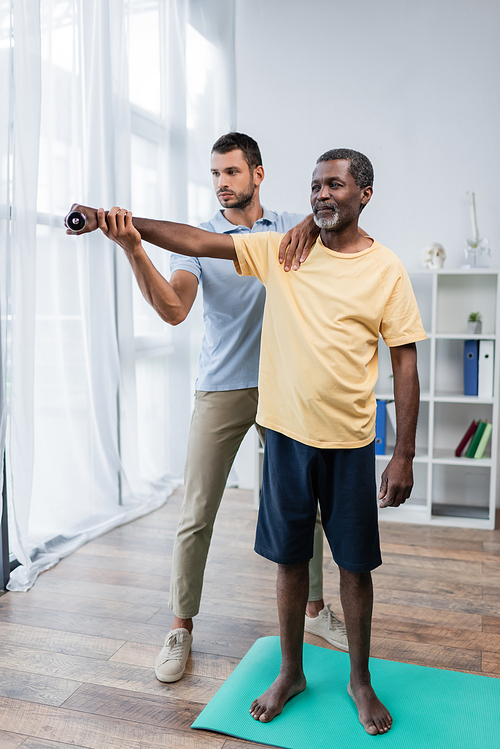 full length view of barefoot african american man exercising with dumbbell near rehabilitologist