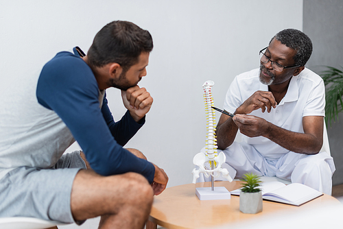 african american physiotherapist pointing at spine model near young man in consulting room