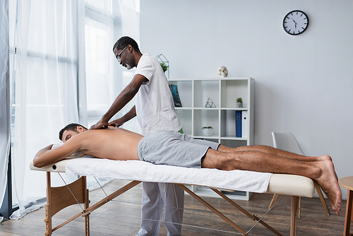 young man lying on massage table during massotherapy by african american rehabilitologist