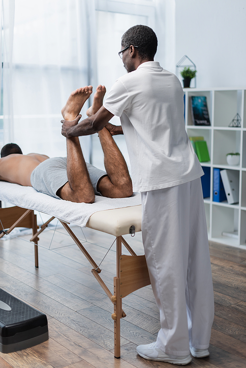 african american rehabilitologist stretching legs of young man on massage table