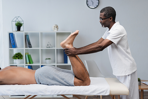side view of african american physiotherapist treating man lying on massage table