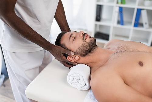young man with closed eyes lying on massage table during rehabilitation treatment by african american masseur