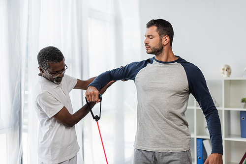 mature african american trainer helping young man exercising with resistance band in rehabilitation center