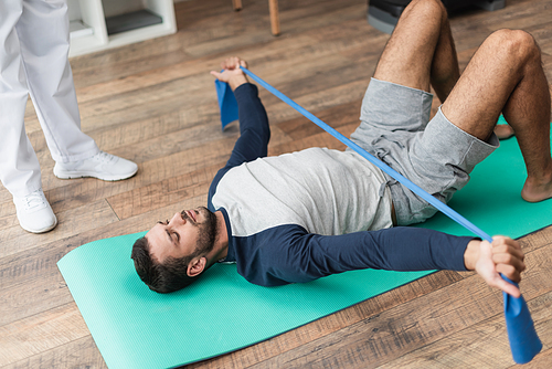 young man lying on fitness mat and training with rubber band near rehabilitologist