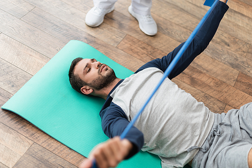 young man working out with rubber band while lying on fitness mat near rehabilitologist