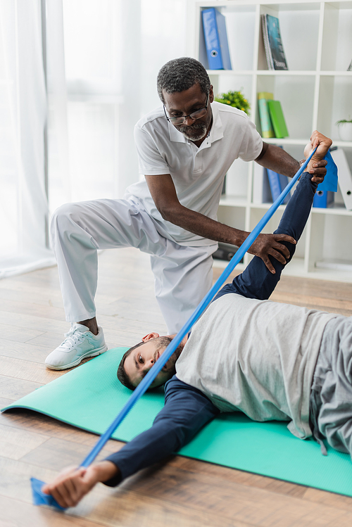 mature african american rehabilitologist helping young man training with rubber band on fitness mat