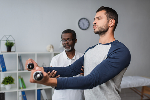 young man exercising with dumbbells near blurred african american rehabilitologist