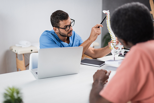 physiotherapist in eyeglasses pointing at spine model near blurred african american patient