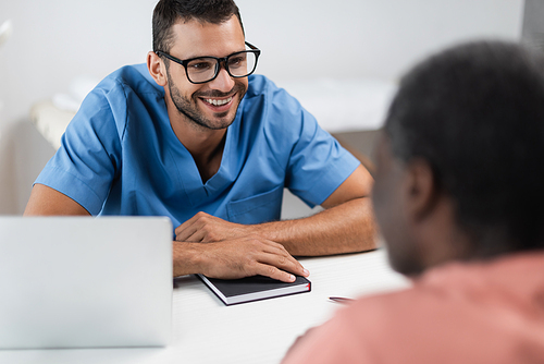 young doctor in eyeglasses smiling near blurred african american patient in consulting room