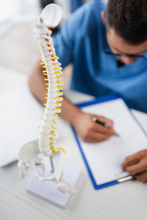selective focus of spine model near blurred rehabilitologist writing prescription on clipboard