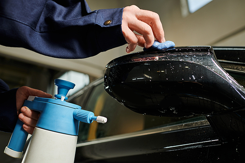 cropped view of hard working serviceman in blue uniform using pulverizer and rag to clean car