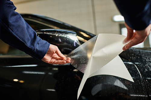 cropped view of hard working man in blue uniform applying protective foil on mirror of black car