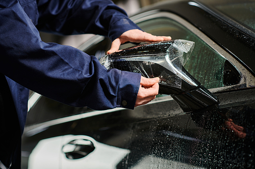 cropped view of professional garage worker in blue uniform applying protective foil on black car