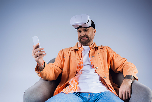 A man immersed in the virtual world, sits in a chair holding a cell phone in a studio setting.