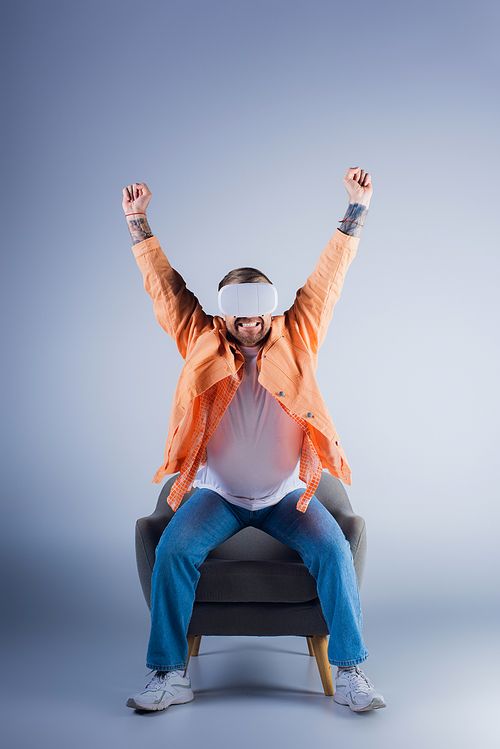 Man in VR headset sits atop chair with arms raised in celebration in a studio setting.