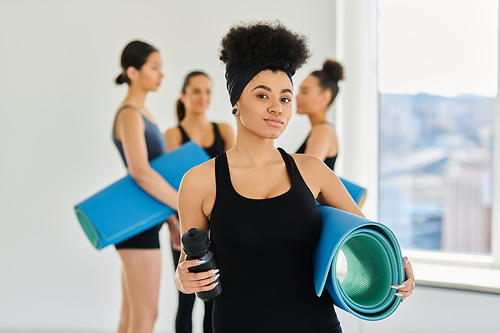 focus on african american woman standing with fitness mat and sports bottle in yoga studio