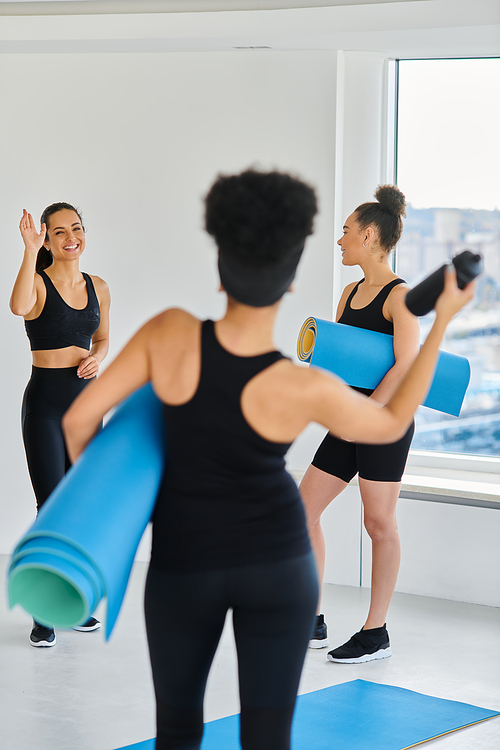 happy brunette woman waving hands at her african american friend with fitness mat in yoga studio