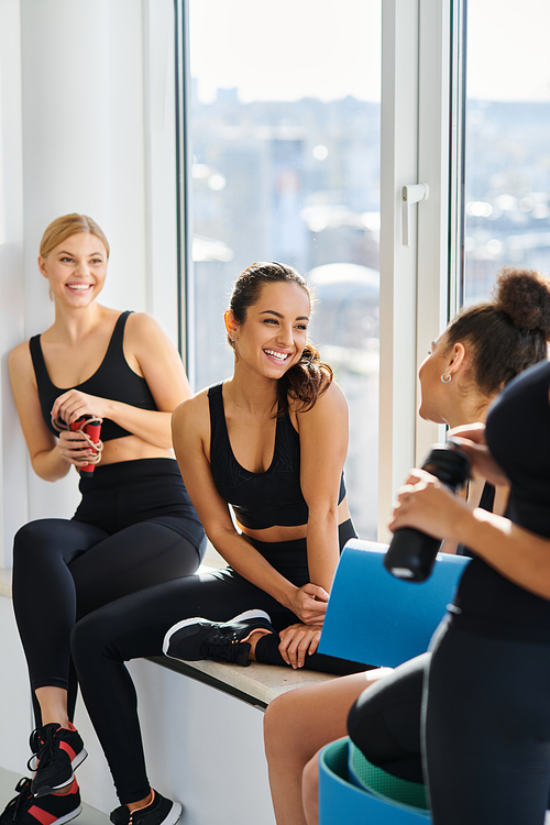 cheerful interracial young women in 20s chatting while sitting next to window in yoga studio