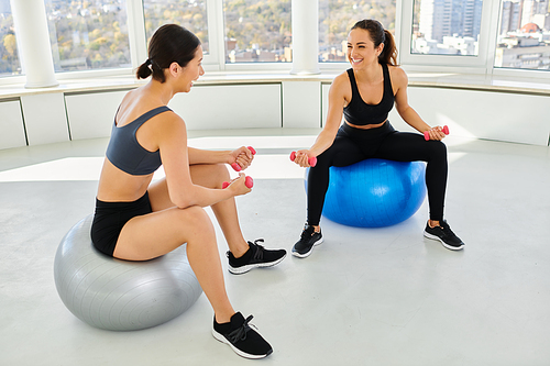 cheerful diverse female friends exercising with dumbbells on fitness balls during pilates class