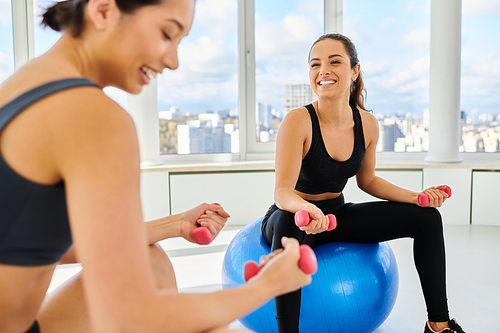 happy interracial female friends exercising with dumbbells on fitness balls during pilates class