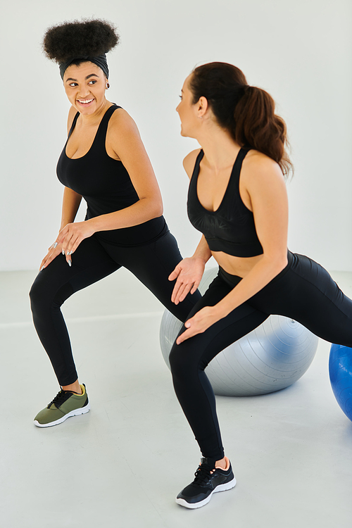 two happy diverse female friends exercising on fitness balls during pilates class, motivation