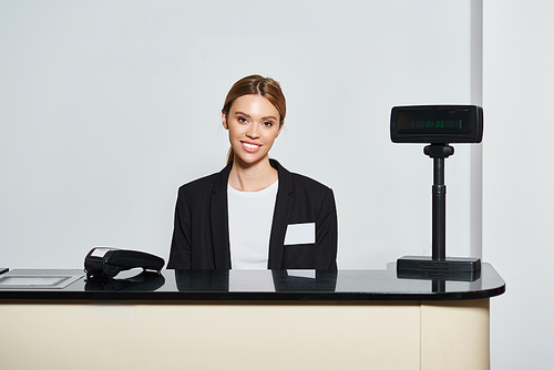 attractive elegant saleswoman in stylish black blazer sitting at counter and looking at camera