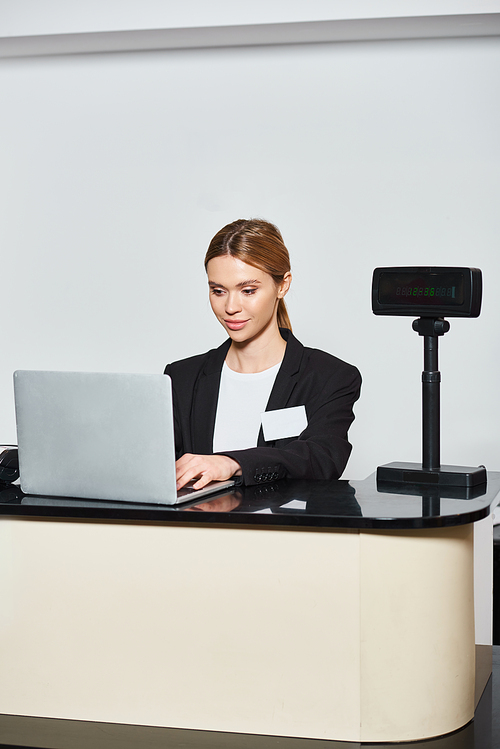 appealing stylish saleswoman in black blazer working on laptop while sitting at counter in store