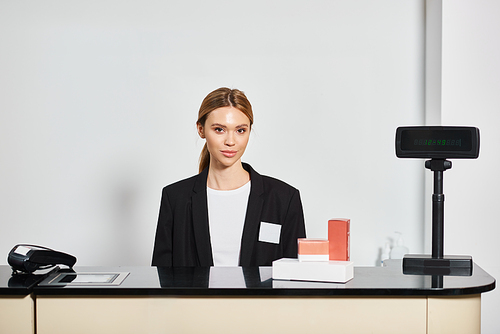 good looking shop assistant in sophisticated black blazer sitting at counter and looking at camera