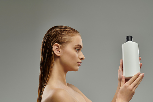 A young woman with wet hair delicately holds a bottle of shampoo in her hand.