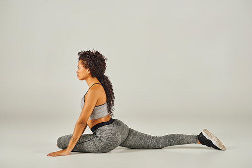 A young African American woman in active wear gracefully stretches in a squat position on a grey background.