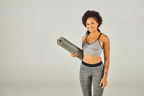 A curly African American sportswoman in activewear holding a yoga mat in a studio with a grey background.