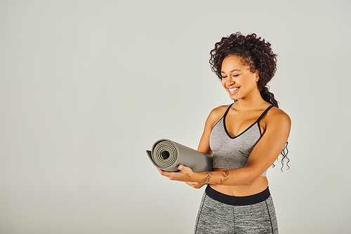 A curly African American sportswoman in athletic attire holds a yoga mat in a studio with a gray backdrop.