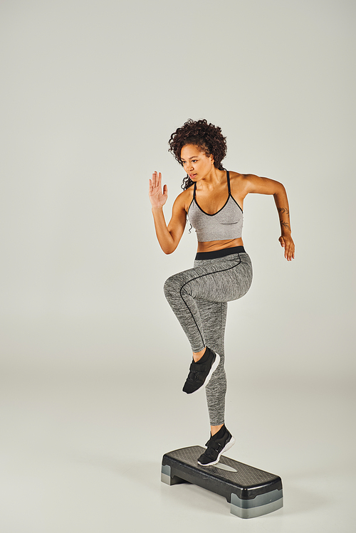 Curly Afro-American sportswoman in activewear gracefully works out on a stepper in a studio with a grey background.