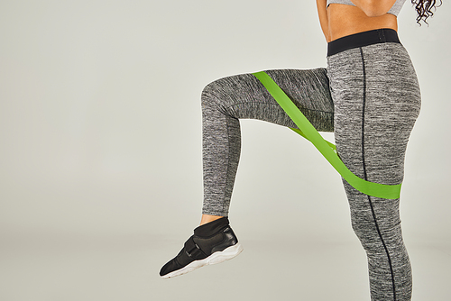 A sportswoman with curly hair wears green band leggings while working out in a studio with a grey background.